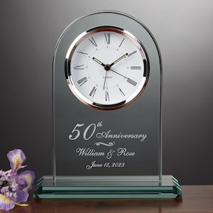 May include: A clear glass clock with a silver face and black hands. The clock has a green base and is engraved with the text "50th Anniversary William & Rose June 15, 2023".