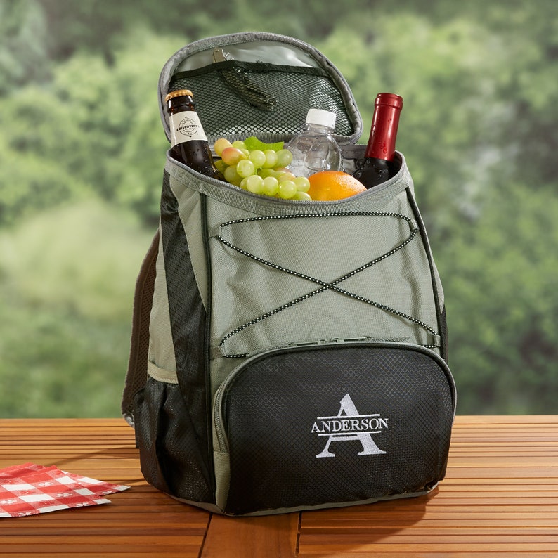 Puede incluir: Una mochila refrigeradora verde y negra con cierre de cremallera en la parte superior y un bolsillo frontal. La mochila tiene una letra "A" bordada personalizada con el nombre "ANDERSON" debajo. La mochila est&aacute; llena de una botella de vino, una botella de cerveza, uvas y una naranja.