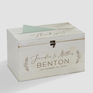 May include: A rectangular white distressed wooden box with a hinged lid and a metal latch. The top features a slot with a light green card. The front is printed with the text "Jennifer & Matthew BENTON SEPTEMBER 24, 2026" and leaf motifs.
