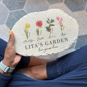 Pu&ograve; includere: Una pietra da giardino personalizzata con una superficie naturale e strutturata. Presenta quattro illustrazioni floreali: un narciso, un papavero, agrifoglio e fiori rosa. Il testo recita "LITA'S GARDEN love grows here" con nomi e date.