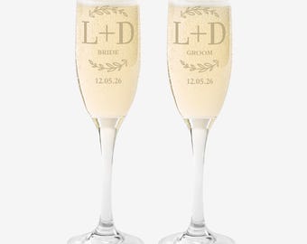 May include: Two champagne flutes filled with a bubbly beverage. The flutes are etched with the letters "L+D", the words "Bride" and "Groom", a sprig design, and the date "12.05.26". The flutes have a long stem and a clear glass base.