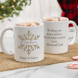 May include: Two white ceramic mugs filled with hot chocolate and marshmallows. One mug features a gold glitter snowflake design with the name "Caroline" and the words "Happy Holidays." The other mug has the message "Wishing you peace and joy this holiday season. Love, Mom and Dad."