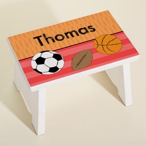 May include: White wooden step stool with a personalized name "Thomas" on a red and orange background with a soccer ball, football, and basketball design.