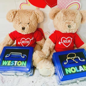 Puede incluir: Dos osos de peluche beige con camisetas rojas con dise&ntilde;os de coraz&oacute;n y nombres, frente a huchas azules. Las huchas tienen los nombres "WESTON" y "NOLAN". El fondo incluye decoraciones del D&iacute;a de San Valent&iacute;n.