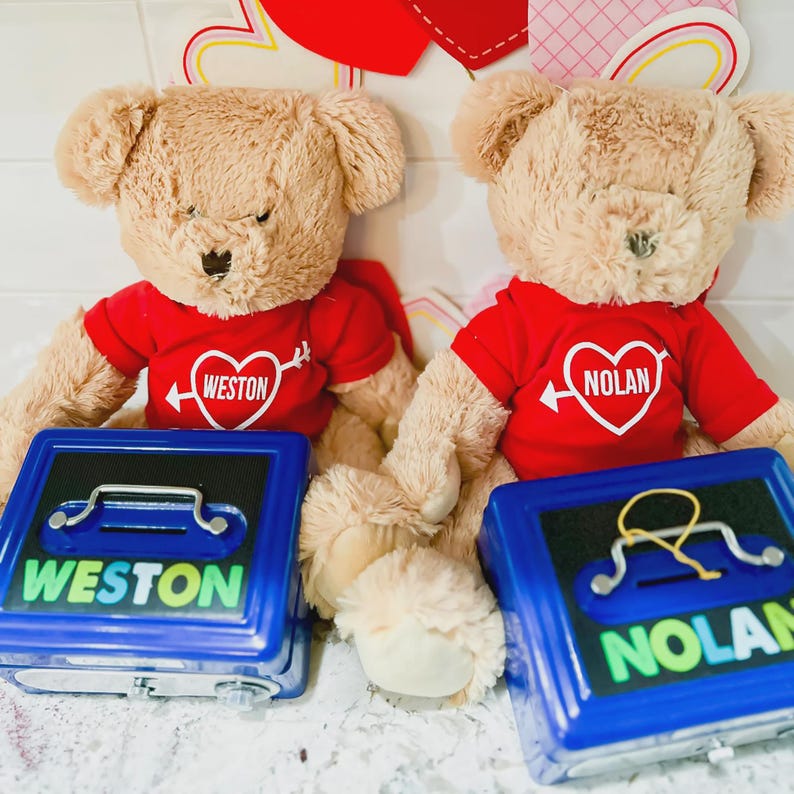 Puede incluir: Dos osos de peluche beige con camisetas rojas con dise&ntilde;os de coraz&oacute;n y nombres, frente a huchas azules. Las huchas tienen los nombres "WESTON" y "NOLAN". El fondo incluye decoraciones del D&iacute;a de San Valent&iacute;n.