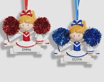 May include: Two hanging ornaments shaped like cheerleaders with blonde hair, one in a red uniform with red pom-poms and the name EMMA, and one in a blue uniform with blue pom-poms and the name OLIVIA. Both figures are in a split position.