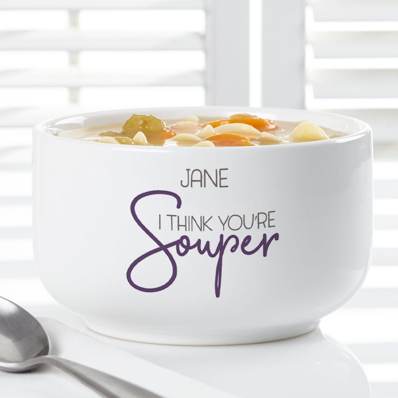 K&ouml;nnte beinhalten: Eine wei&szlig;e Keramik-Suppensch&uuml;ssel gef&uuml;llt mit Suppe, mit Karotten, Nudeln und Gem&uuml;se. Die Sch&uuml;ssel tr&auml;gt den Text "JANE I THINK YOU'RE Souper" in lila Schrift. Ein silberner L&ouml;ffel liegt neben der Sch&uuml;ssel.