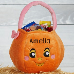 May include: Orange plush pumpkin-shaped basket with a pink handle and bow, filled with candy. The basket has a smiling face and the name "Amelia" embroidered on it. A festive container for treats.