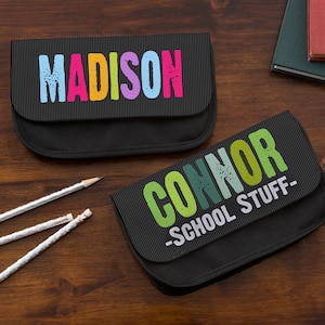 May include: Two black rectangular pencil cases with textured flaps. One case features the name MADISON in multicolored block letters. The other case features the name CONNOR and the text -SCHOOL STUFF- in green and white distressed lettering.