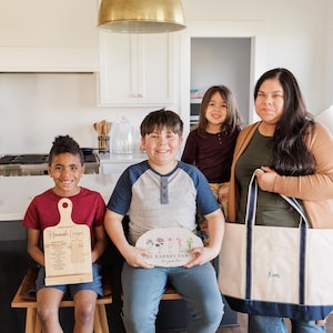 Pu&ograve; includere: Una famiglia posa in una cucina, tenendo oggetti personalizzati. Sono visibili un tagliere in legno con testo di ricetta, una targa floreale con il nome della famiglia e una grande borsa tote con un nome ricamato.