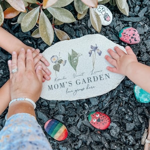 Pu&ograve; includere: Una targa da giardino in pietra con le parole "MOM'S GARDEN" e "love grows here" stampate sopra. La targa &egrave; circondata da sassi dipinti e mani. La targa presenta illustrazioni floreali e nomi.