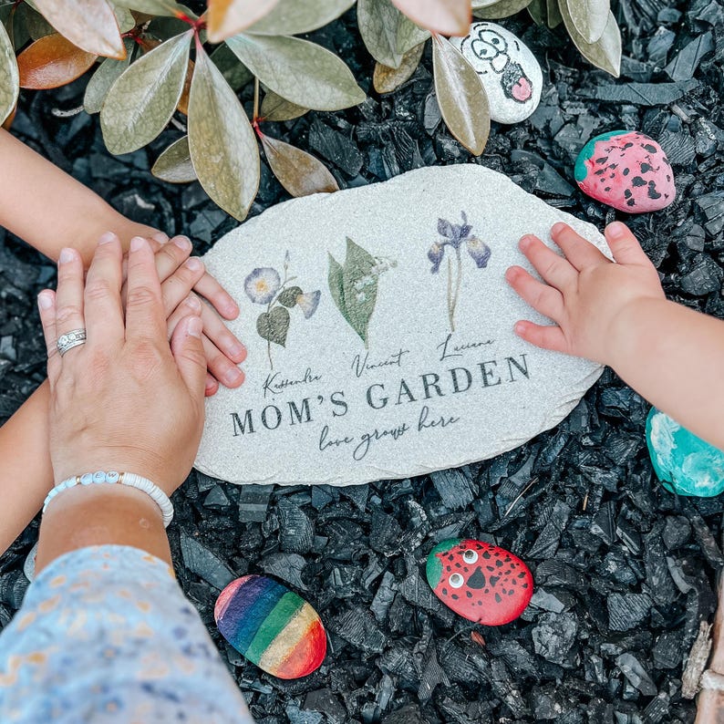 Pu&ograve; includere: Una targa da giardino in pietra con le parole "MOM'S GARDEN" e "love grows here" stampate sopra. La targa &egrave; circondata da sassi dipinti e mani. La targa presenta illustrazioni floreali e nomi.