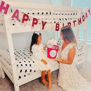 Puede incluir: Una litera blanca con una colcha de lunares blancos y negros y una pancarta que dice "HAPPY BIRTHDAY". Una cubeta rosa con "Happy Birthday" es sostenida por una mujer y entregada a una ni&ntilde;a peque&ntilde;a con un vestido blanco.