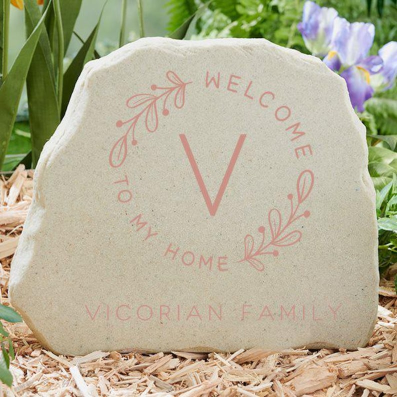 May include: A beige stone garden marker with the words "WELCOME TO MY HOME" and "VICTORIAN FAMILY" in pink. A pink "V" and floral wreath design are in the centre. The stone is set in a bed of wood chips, with greenery and flowers in the background.