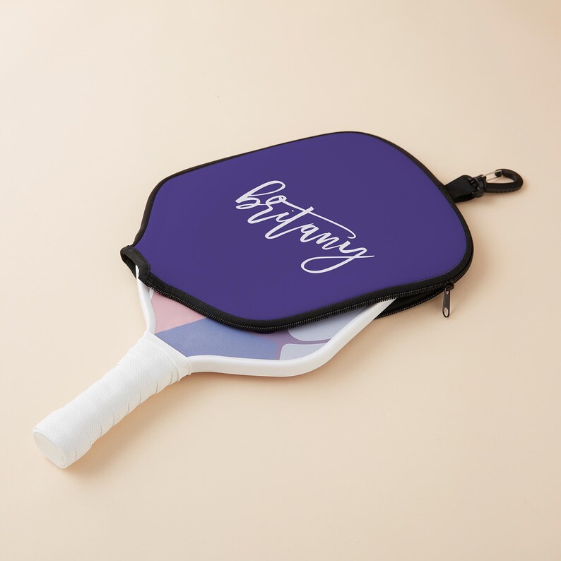 May include: A purple pickleball paddle cover with a zipper closure and a white strap with a clip. The cover has the name "Brittany" written in white cursive lettering.