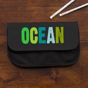 May include: A black pencil case with a vertical pinstripe pattern on the flap. The word OCEAN is printed in large, distressed, multi-colored letters including lime green, teal, and light blue. Two white pencils are positioned above the case.