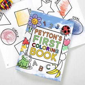 May include: A blue and white coloring book with the title "Peyton's First Coloring Book" and a colorful illustration of a frog, a banana, a ladybug, and a flower. The book also features the letters A, B, and C.