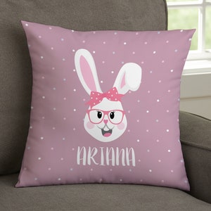 May include: A decorative square pillow with a pink background and white polka dots. The pillow features a cartoon bunny face with pink glasses, a bow, and the name "Ariana" in white letters. The bunny has white ears and a smiling face.