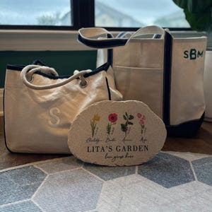 Pu&ograve; includere: Tre borse di tela e una pietra decorativa. Le borse sono bianco sporco con finiture e manici blu navy. Una borsa ha la lettera "S", e un'altra le iniziali "SBM". La pietra recita "LITA'S GARDEN love grows here" con illustrazioni floreali.