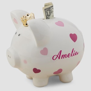 May include: White ceramic piggy bank with pink hearts and the name "Amelia" in a cursive font. The piggy bank has a gold crown and a slot for inserting money. A dollar bill is visible in the slot.