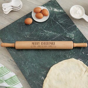 Puede incluir: Un rodillo de madera con el texto "Merry Christmas The Anderson Family" grabado en &eacute;l. El rodillo est&aacute; sobre una superficie de m&aacute;rmol verde con un taz&oacute;n de huevos y una masa.