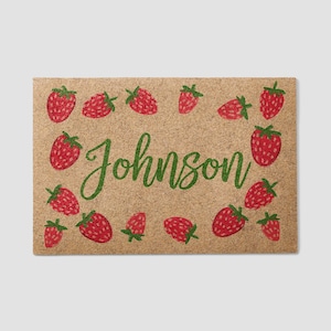 May include: A rectangular tan coir doormat featuring a repeating pattern of red strawberries with green stems around the perimeter. The name Johnson is printed in the center in a green cursive script font across the middle of the mat.
