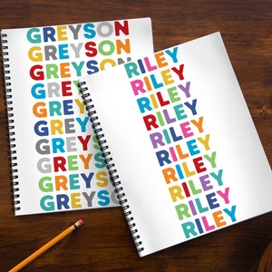 May include: Two spiral bound notebooks with colorful names repeated on each page. The notebook on the left has the name Greyson repeated in various colors. The notebook on the right has the name Riley repeated in various colors.