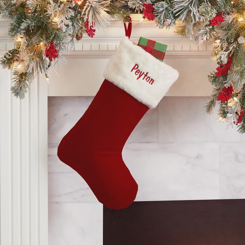 May include: A deep red Christmas stocking with a white faux fur cuff featuring the embroidered name Peyton in red thread. A small gift box wrapped in green and red paper is tucked inside the top of the stocking, which hangs from a red loop.