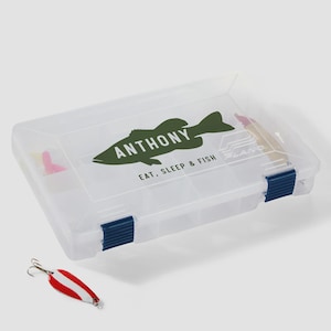 May include: A clear plastic tackle box with two blue latches, featuring a green fish graphic with the text ANTHONY and EAT, SLEEP & FISH printed on the lid. A red and white metal fishing spoon lure with a silver hook lies in front of the box.