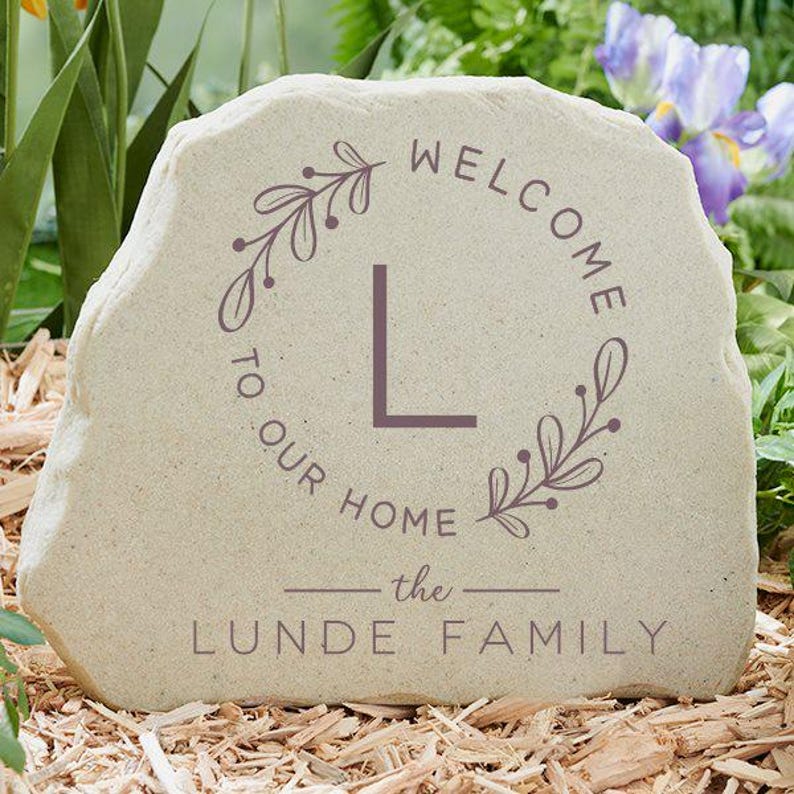 May include: A beige stone with the words "WELCOME TO OUR HOME" and "the LUNDE FAMILY" in dark purple lettering. A purple floral wreath surrounds the letter "L". The stone is sitting on a bed of wood chips.