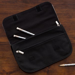 May include: A black fabric pencil case with a fold-over flap secured by hook-and-loop fasteners. The interior features a zippered compartment and an open pocket containing two silver-patterned pencils, with a third pencil resting outside.