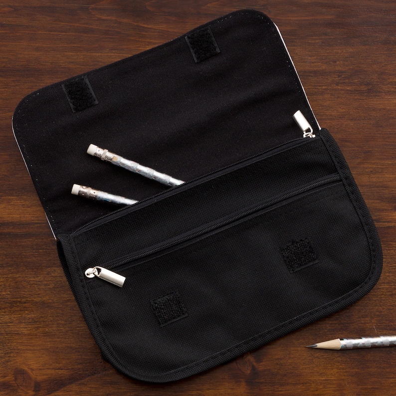 May include: A black fabric pencil case with a fold-over flap secured by hook-and-loop fasteners. The interior features a zippered compartment and an open pocket containing two silver-patterned pencils, with a third pencil resting outside.