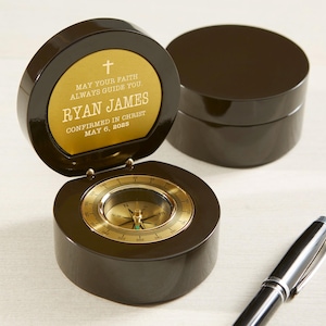 May include: A dark brown compass with a gold-colored face and a lid that opens to reveal a personalized message. The message reads "MAY YOUR FAITH ALWAYS GUIDE YOU. RYAN JAMES CONFIRMED IN CHRIST MAY 6, 2025." A black pen rests nearby.