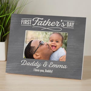 May include: Gray wooden picture frame with the text "First Father's Day" and "Daddy & Emma". The frame holds a photo of a father kissing a baby. The text "I love you, Daddy!" is also visible.