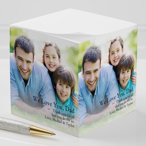 May include: A white cube-shaped notepad with a photo of a father and his two children. The notepad has the text "We Love You, Dad" and "Thank you for all that you do for us. Love, Michael & Taylor" printed on it.