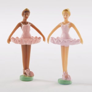 May include: Two plastic ballerina figurines, one with brown skin and one with fair skin, both wearing pink tutus and standing on green bases.