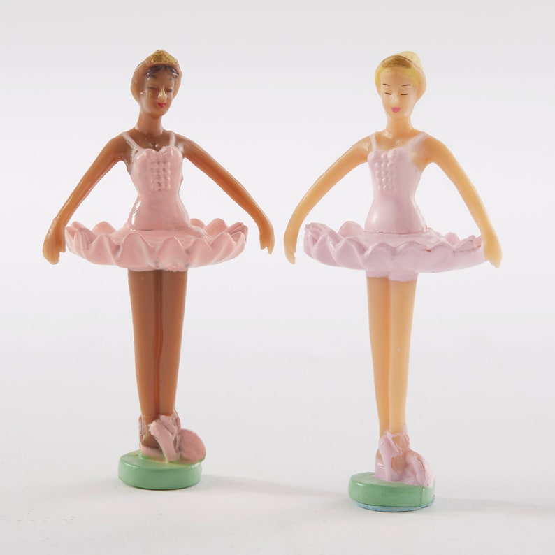 May include: Two plastic ballerina figurines, one with brown skin and one with fair skin, both wearing pink tutus and standing on green bases.