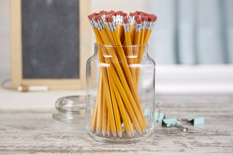 May include: A glass jar filled with yellow pencils with red erasers. The jar is sitting on a wooden surface.