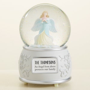 May include: A white snow globe with a silver base and a small angel figurine inside. The base has the text "THE THOMPSONS An Angel from above protects our family".