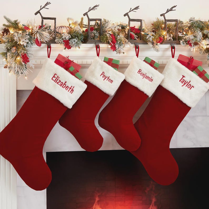 May include: Four deep red Christmas stockings with white faux fur cuffs, each embroidered with a name: Elizabeth, Peyton, Benjamin, and Taylor. Small wrapped gifts are tucked into the top of each stocking, which hang from metal reindeer hooks.