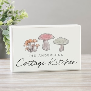 May include: A rectangular wooden sign with a white frame. The sign features watercolor illustrations of mushrooms in shades of brown, pink, and green. The text reads "THE ANDERSONS Cottage Kitchen."