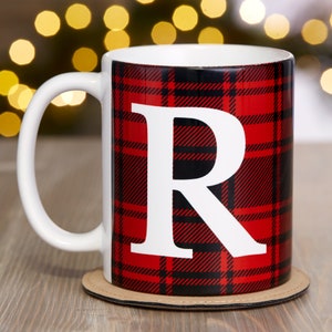 Puede incluir: Una taza de cer&aacute;mica blanca con asa negra. La taza tiene un patr&oacute;n de cuadros rojos y negros con una letra "R" blanca en el centro. La taza est&aacute; sobre un posavasos marr&oacute;n.