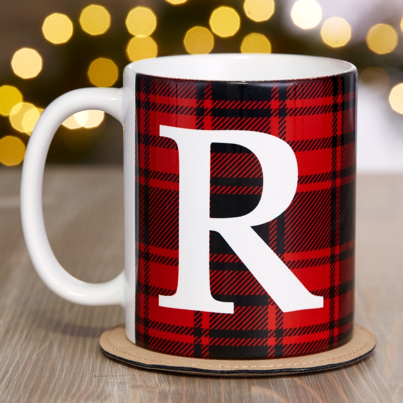 Puede incluir: Una taza de cer&aacute;mica blanca con asa negra. La taza tiene un patr&oacute;n de cuadros rojos y negros con una letra "R" blanca en el centro. La taza est&aacute; sobre un posavasos marr&oacute;n.
