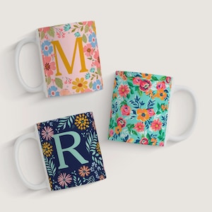 May include: Three white ceramic mugs with floral patterns and initial letters. The mugs are on a white background. The first mug has a pink background with yellow, orange, and pink flowers and the letter "M" in gold. The second mug has a blue background with pink, orange, and yellow flowers. The third mug has a dark blue background with pink, yellow, and orange flowers and the letter "R" in light blue.