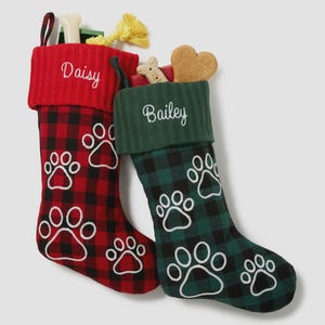 May include: Two Christmas stockings for pets, one red and one green, with paw print designs. The red stocking has the name "Daisy" embroidered on the cuff, and the green stocking has "Bailey". Each stocking is filled with dog toys and treats.
