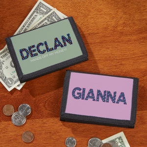 May include: Two personalized wallets with the names "Declan" and "Gianna" printed on them. The "Declan" wallet has a green background with blue and red patterned letters, and the text "HANDS OFF MY MONEY!". The "Gianna" wallet has a pink background.
