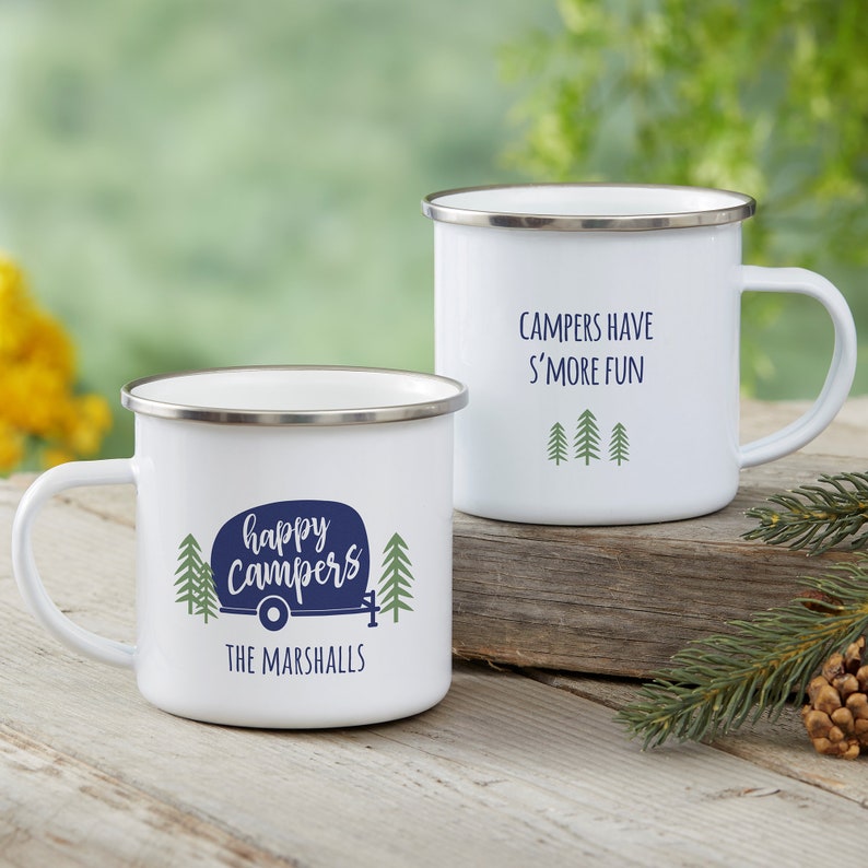 May include: Two white enamel mugs with silver rims. One mug features a blue camper graphic, green pine trees, and the text "happy campers THE MARSHALLS". The other mug displays the text "CAMPERS HAVE S'MORE FUN" above three green pine trees.