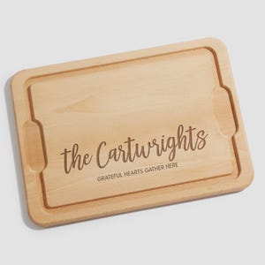 May include: A rectangular light-colored wooden serving board with integrated side handles and a juice groove. The board is engraved with the text "the Cartwrights" in a script font and "GRATEFUL HEARTS GATHER HERE" in a sans-serif font.