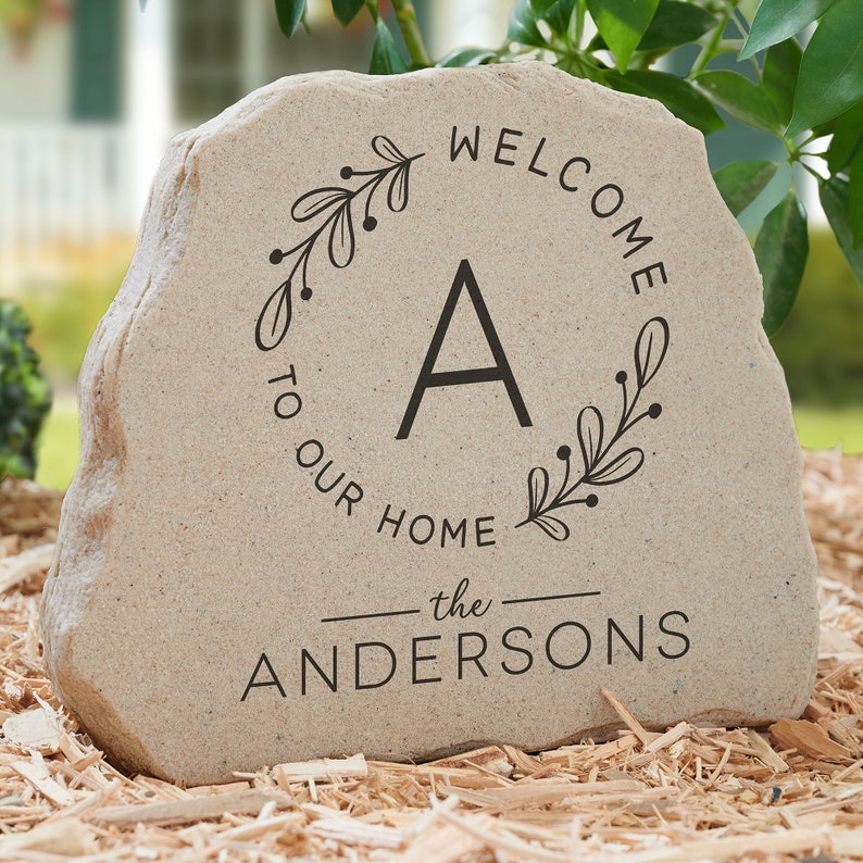 May include: A beige stone with the words "WELCOME TO OUR HOME" and "the ANDERSONS" in black lettering. A black letter "A" is centered within a floral wreath. The stone is on a bed of wood chips.