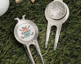 May include: Two silver golf ball divot tools. One has a rainbow circle with the text "Best Daddy By Par" and the other has a dimpled golf ball design.
